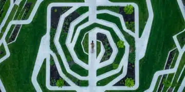 An aerial drone view of a person walking on winding geometric pathways in an urban park, symbolizing the evolving workforce and complex hybrid work models. The pathways are concrete, bordered by green lawns, under an overcast sky.