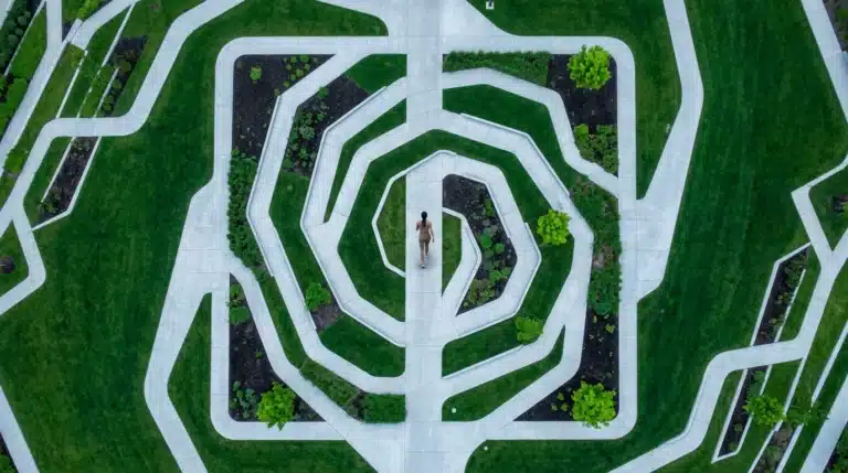 An aerial drone view of a person walking on winding geometric pathways in an urban park, symbolizing the evolving workforce and complex hybrid work models. The pathways are concrete, bordered by green lawns, under an overcast sky.